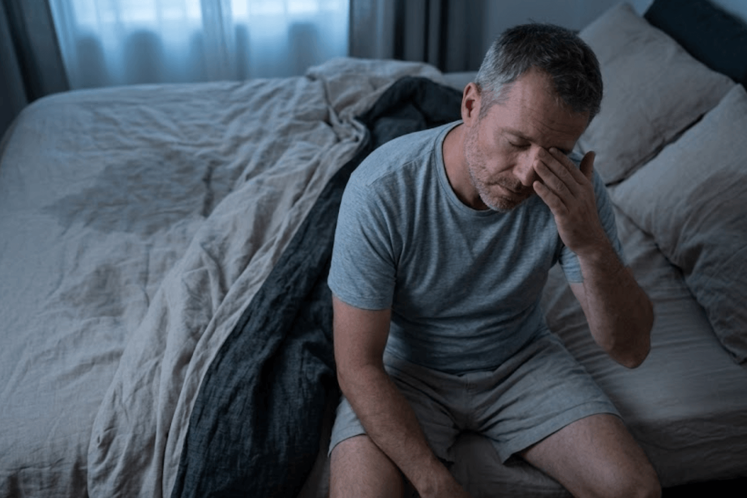 Man sitting on the edge of a bed looking exhausted from sleep deprivation and night sweats.